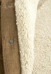 Brown corduroy fabric with a ribbed texture pairs with soft, cream-colored fleece. A silver button accent is visible near the seam.