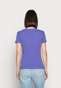 Nainen, jolla on suorat tummat hiukset, pukeutuneena lyhythihaiseen violettiin t-paitaan ja vaaleansinisiin farkkuihin, kääntynyt pois yksinkertaista taustaa vasten.