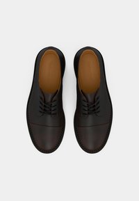 Sapatos de couro pretos com um acabamento texturizado, bico redondo, design com atacadores e detalhes castanhos. Apresentam uma sola robusta e costura visível.