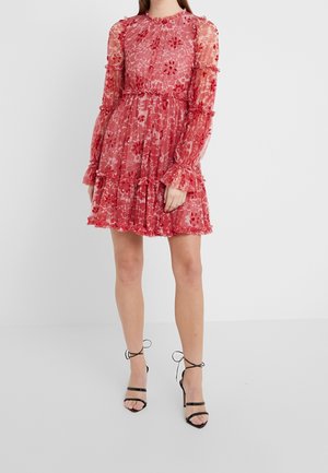 Kobieta w czerwonej, kwiecistej, przezroczystej sukience z długim rękawem, sięgającej do kolan, z falbanami i czarnych sandałkach na wysokim obcasie z paskami.