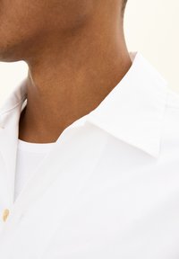 Een witte, colbertachtige shirt zichtbaar vanaf de zijkant, met een gladde textuur en een enkele knoopdetail op de kraag. Daaronder een witte t-shirt.