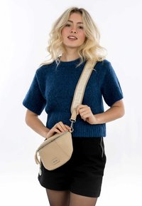 Borsa a tracolla in pelle beige con forma triangolare, chiusura con zip e tracolla rimovibile. Il maglione è di maglia blu con maniche corte.