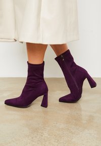 Botas de tobillo de ante morado con puntera afilada, tacón alto en bloque y cierre de cremallera en el lado exterior. Textura suave con detalles de costura sutil.