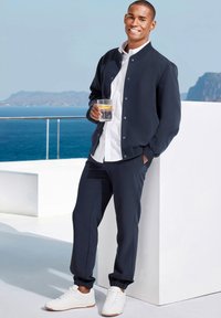 Navy bomber jacket over a white shirt, paired with matching navy joggers and white sneakers. Holding a glass with a lemon slice.