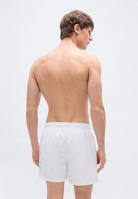 Λευκά σορτς για κολύμπι με ελαστική μέση, featuring μικρό λογότυπο στο αριστερό μέρος. Λείο, ελαφρύ υλικό. Πλάτη χωρίς ρούχα.