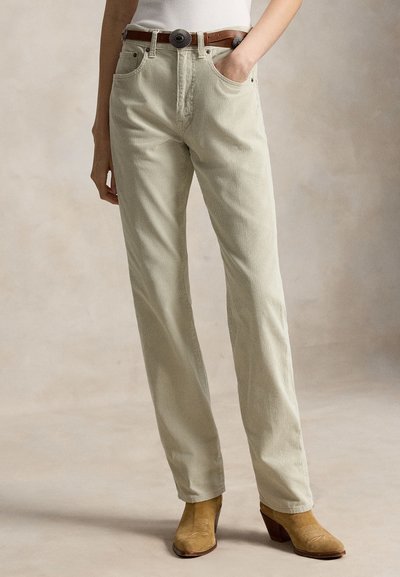 Pantalon en velours côtelé beige à coupe droite, porté avec une ceinture marron et des bottines marron clair, une main dans la poche, arrière-plan neutre.