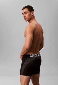 Calzoncillos bóxer negros de Calvin Klein con un panel lateral texturizado y una cinturilla ancha elástica con la marca en blanco. Modelo de espaldas.