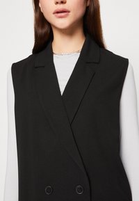 Chaleco negro sin mangas con un diseño de doble botonadura, solapas marcadas y dos botones. Lleva encima una camisa blanca de manga larga. Textura de tela suave.