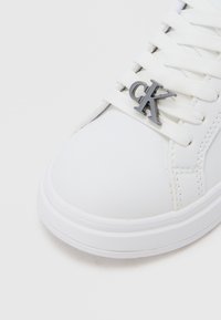 Λευκό αθλητικό παπούτσι με λευκά κορδόνια που φέρουν λεπτομέρεια με ασημένιο λογότυπο "CK" κοντά στην περιοχή των δακτύλων.