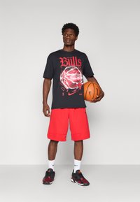 T-shirt preta com gráfico vermelho dos "Chicago Bulls", combinada com calções vermelhos. Meias brancas e sapatos pretos com detalhes vermelhos. Segurando uma bola de basquete.