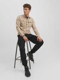 Beige plaid button-up shirt with two chest pockets, paired with black cargo pants and black athletic shoes, seated on a metal stool.