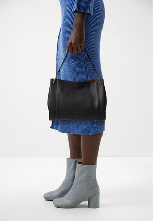 Kobieta ubrana w niebieską sukienkę z prążkowanego materiału i jasnoszare botki do kostki, trzymająca czarną skórzaną torebkę z cienkimi rączkami po boku.