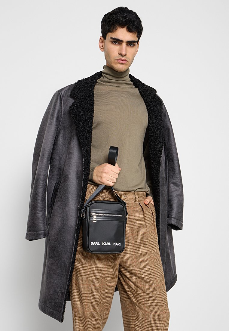 Bolso bandolera cuadrado negro con cierre de cremallera y detalle de logo, hecho de material liso, llevado por una modelo con abrigo gris y pantalones estampados.