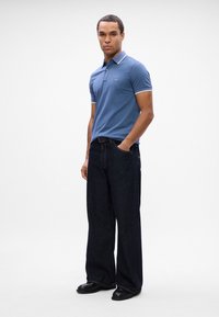 Polo bleu clair avec un col blanc contrastant, associé à un jean large bleu foncé et des chaussures noires, se tenant devant un fond clair.