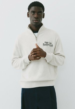Jeune homme à la peau foncée portant un pull léger avec fermeture éclair et le texte "High Line Running Club", debout devant un fond blanc uni.