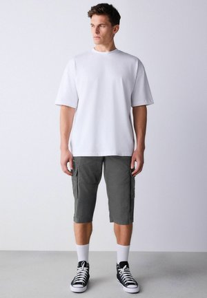 Vyras dėvi per didelį baltą marškinėlį, tamsiai pilkas krovinines šortus, baltas aukštas kojines ir juodus aukšto viršaus sportbačius, stovintis žvelgdamas į priekį.