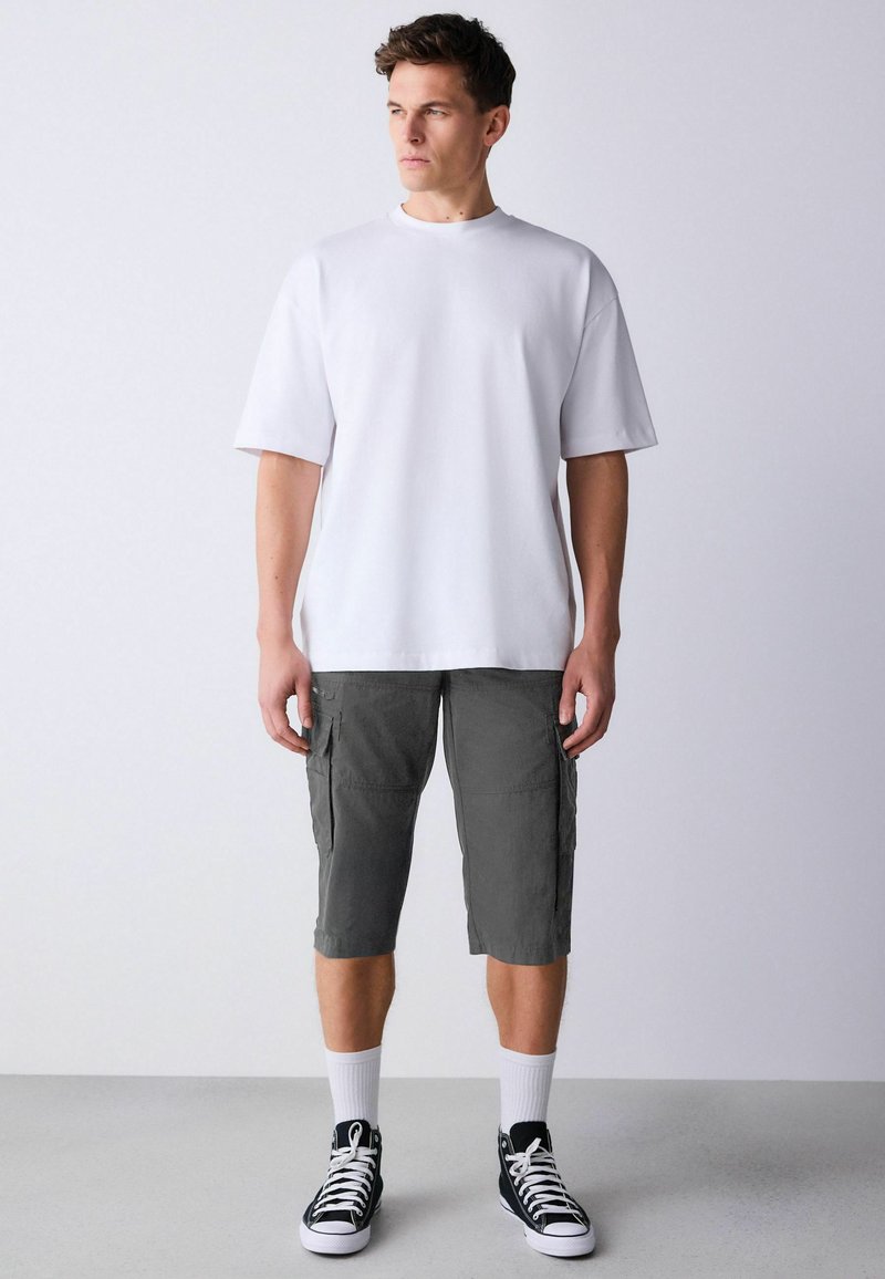 Homme portant un t-shirt blanc surdimensionné, un short cargo gris foncé, des chaussettes blanches mi-mollet et des baskets montantes noires, debout face à l'avant.
