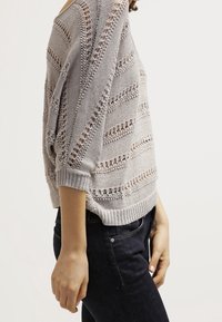 Pull en tricot gris avec des motifs à mailles ouvertes, doté d'une coupe ample et de poignets côtelés, associé à un jean noir.