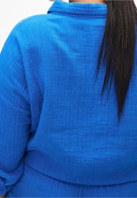 Blue textured garment with a collar and rolled sleeves, featuring a relaxed fit and visible stitching patterns throughout the fabric.