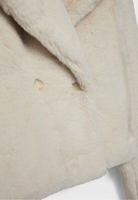 Nærbillede af beige imiteret pelsfrakke stof med to cremefarvede knapper og en foldet revers på frontpanelet.