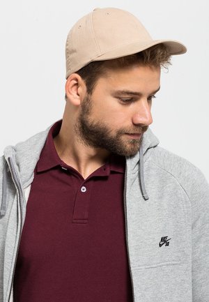 Sivi zip-up hoodie preko tamnocrvene polo majice, s bež bejzbol kapom. Hoodie ima Nike logo na prsima i vezicu. Obični uzorci.