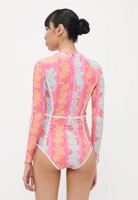 Maillot de bain à manches longues en rose avec des motifs floraux en orange et bleu, accents blancs, et fermeture éclair au dos. Design texturé et ajusté.