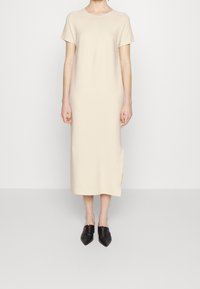 Abito beige al ginocchio con maniche corte, scollatura rotonda e spacchi laterali, realizzato in tessuto morbido; abbinato a scarpe nere con punta a punta.