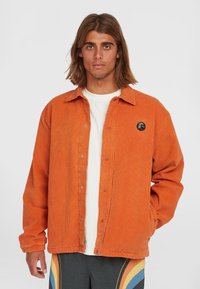 Giacca di corduroy arancione con bottone anteriore, colletto a punta e patch con logo nero. Il modello indossa una maglietta bianca e pantaloni a fantasia.