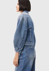 Giacca di jeans con una forma strutturata, maniche lunghe e un colore blu sbiadito. Presenta un colletto alto e tasche posteriori, mostrando un design classico.