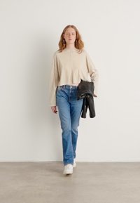 Beige kort genser med sidesømmer, blå rette jeans, og hvite sneakers. Holder en svart skinnjakke. Nøytral bakgrunn.