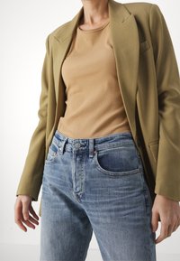 Oliwkowa marynarka na jasnobrązowym, prążkowanym topie, w połączeniu z jasnoniebieskimi dżinsami z metalowymi nitami i zapięciem na guzik.