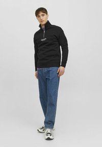Felpa nera con zip e colletto alto, caratterizzata da testo del logo bianco, abbinata a jeans blu dal taglio comodo e scarpe da ginnastica multicolori.