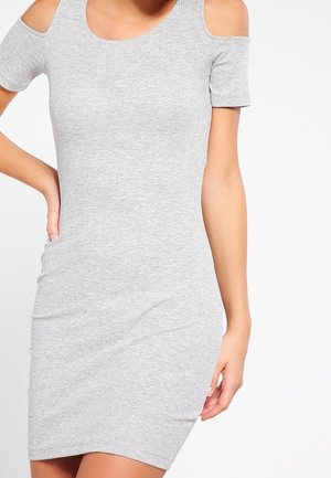 Kobieta ubrana w dopasowaną, szarą sukienkę z krótkim rękawem i wycięciami na ramionach, stojąca z jedną ręką na biodrze na białym tle.