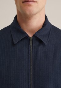 Temno modra zip-up jakna s ovratnikom, ki ima subtilen vertikalni črtast vzorec, teksturirano tkanino in kovinske zadrge.