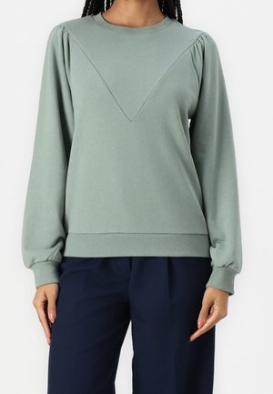 Kvinde iført en lysegrøn langærmet trøje med en V-formet søm og marineblå bukser mod en ensfarvet baggrund.