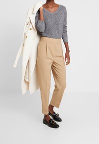 Grauer strukturierter Pullover, beige plissierte Hosen und schwarze Slipper mit goldenen Hardware-Details. Ein cremefarbener Mantel über dem Arm.