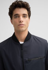 Marineblauer Bomberjacke mit Reißverschluss vorne, geripptem Kragen und einer eleganten Textur; verfügt über eine horizontale Reißverschlusstasche mit Logo-Akzent.