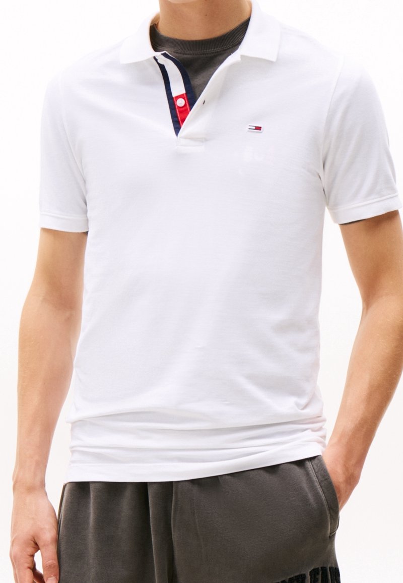 Vit pikétröja i bomull med korta ärmar, med en kontrasterande randig krage och en liten logotyp på bröstet.