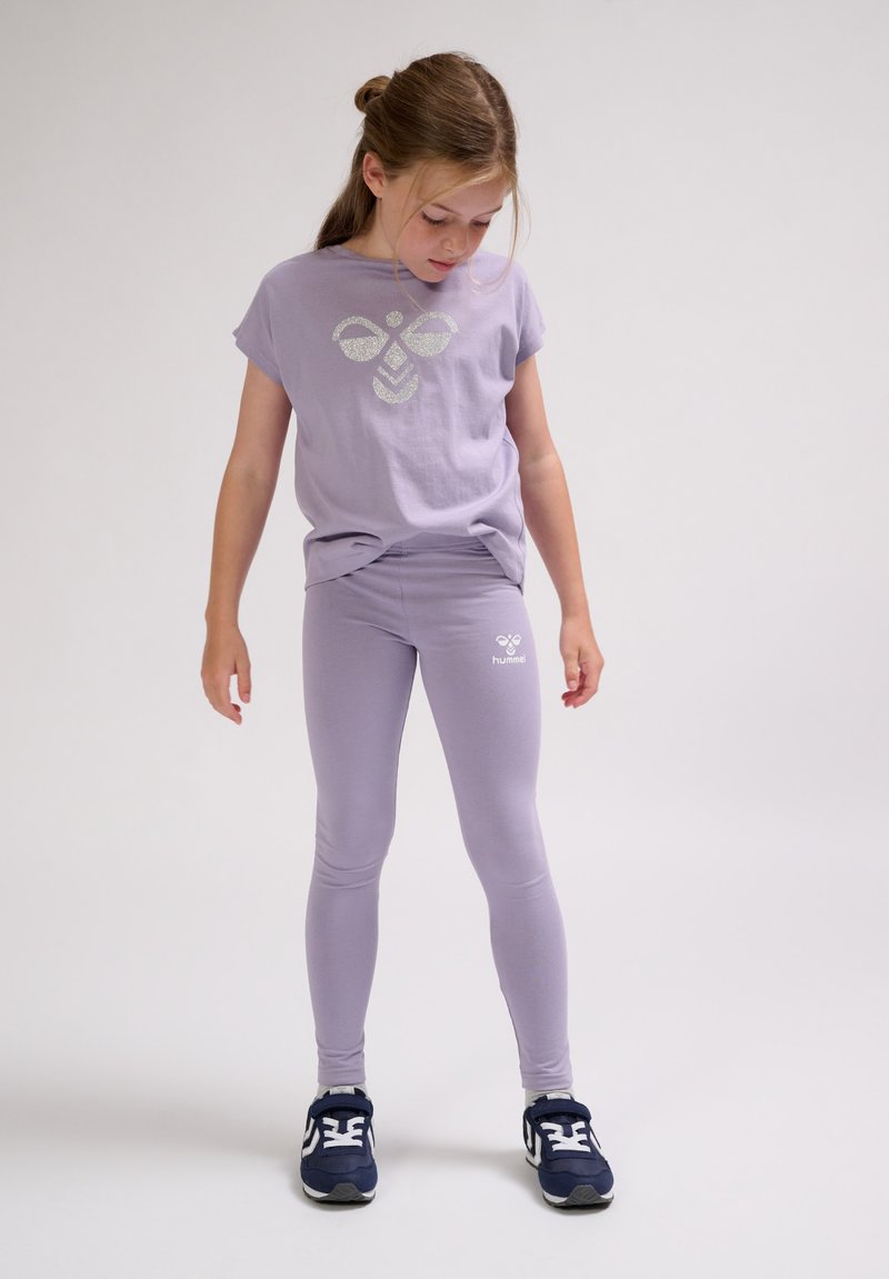 Fille en t-shirt lavande avec logo abeille et leggings assortis, regardant vers le bas, debout devant un fond clair uni avec des baskets bleu marine.