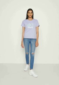 T-shirt lilás claro com o logótipo "Converse" em branco na frente, combinado com jeans skinny azuis rasgados e ténis brancos.