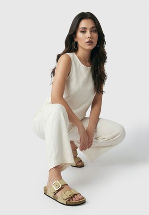 Vrouw met lang donker haar die een mouwloze beige top draagt, bijpassende wijdvallende broek, gouden oorringen en beige sandalen met grote gespen.