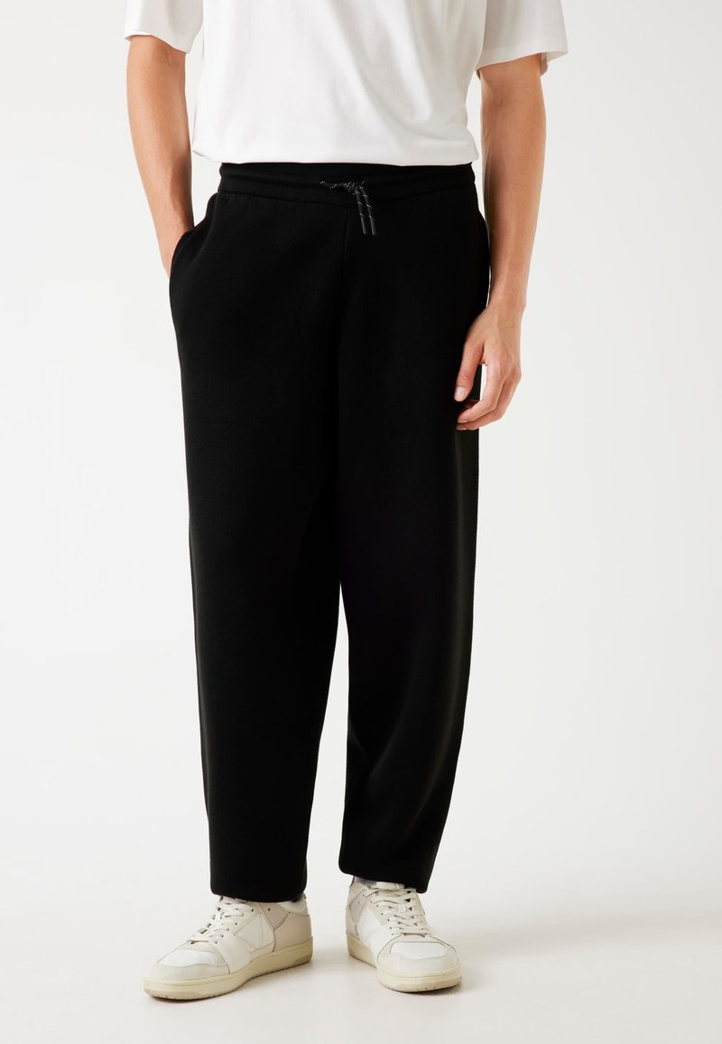 Zwarte joggingbroeken van zachte stof, met een drawstring tailleband, zijzakken en een relaxed fit. Gecombineerd met witte sneakers.