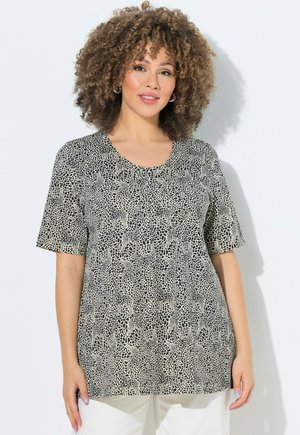 Vrouw met krullend haar met een zwart-wit abstract patroon kort shirt en een witte broek, staand tegen een witte achtergrond.