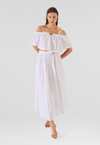 Balts of-the-shoulder krūšturis ar frillētiem piedurknēm un balta maksi svārki ar elastīgu jostasvietu, ar plūstošu auduma tekstūru.