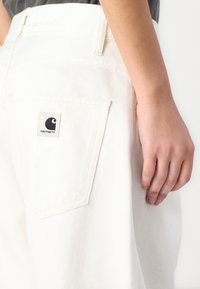 Bílé džínové kalhoty s klasickým střihem, vybavené zadní kapsou a černou nášivkou Carhartt. Tkanina má jemnou strukturu.