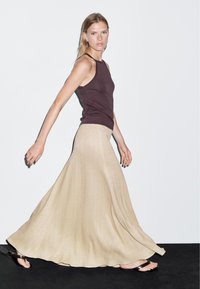 Top marrone senza maniche abbinato a una lunga gonna beige fluente con motivo a spina di pesce. L'outfit ha una texture liscia e un design minimalista.