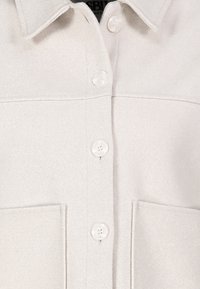 Beige overhemddesign met een kraag, twee voorzakken en een subtiele textuur. Voorzien van ronde witte knopen en een rechte snit.