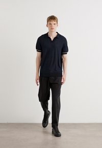 Polo-shirt marine avec un col en V et des manches courtes, présentant des accents rayés blancs. Associé à un pantalon noir ample et des baskets sombres.