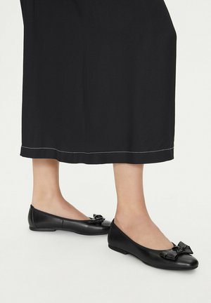 Piedi con ballerine in pelle nera con fiocchi, posati su una superficie chiara sotto una gonna nera.