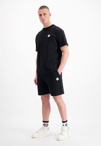 Zwart t-shirt met korte mouwen en logo, gecombineerd met zwarte shorts met zakken en contrasterende witte branding. Witte sneakers en gestreepte sokken maken de look af.